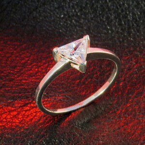 May include: A silver ring with a triangular clear gemstone set in the centre. The ring is on a red leather background.