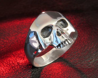 May include: A silver skull ring with a detailed design. The ring is on a red leather surface.