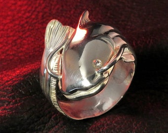 May include: A silver ring featuring a detailed catfish design. The catfish is curled around the band of the ring, with its tail wrapping around the front of the ring.