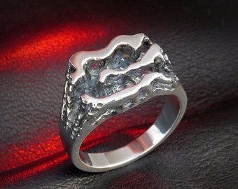 Può includere: Un anello d'argento con un design strutturato e organico. L'anello presenta una superficie ruvida e irregolare con un motivo a onde fluenti. La fascia è liscia e lucida, in contrasto con la parte superiore strutturata. L'anello è posto su uno sfondo scuro con una luce rossa.