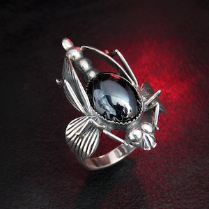 May include: A silver ring with a black gemstone in the shape of a fly. The ring is detailed with intricate wings and a textured body.