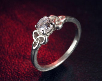 Könnte beinhalten: Ein silberner Ring mit einem klaren, runden Edelstein. Der Ring hat ein keltisches Knotendesign am Band und um die Edelsteinfassung. Der Ring ist vor einem dunkelroten Hintergrund platziert.
