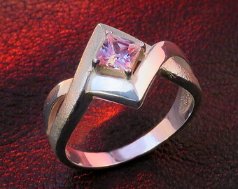 May include: A silver ring with a pink square-shaped gemstone set in the center. The ring has a unique, twisted design.