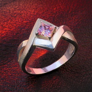 May include: A silver ring with a pink square-shaped gemstone set in the center. The ring has a unique, twisted design.