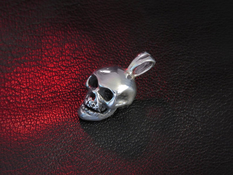 Angled side view of a 0.55″ × 0.35″ sterling silver skull pendant on pebbled black leather, backlit with warm red tones. This perspective highlights the skull’s smooth, rounded contours, crisp undercut around the nasal cavity, and the bail loop’s elegant taper. The interplay of shadows and highlights emphasizes the hand-finished texture and the subtle variations that mark each piece as uniquely made-to-order.