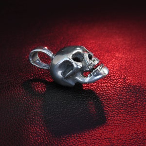 Close-up detail of the bail loop and hallmark on a tiny sterling silver skull pendant set against a textured black leather background. The macro view reveals the precise polishing work around the loop attachment and the clear “925” stamp. Fine surface striations and subtle tool-marks speak to the hand-finished nature of the metal, while the skull’s jawline and teeth are sharply defined.