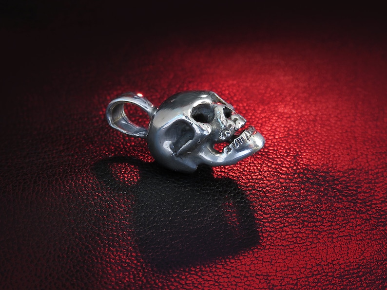 Close-up detail of the bail loop and hallmark on a tiny sterling silver skull pendant set against a textured black leather background. The macro view reveals the precise polishing work around the loop attachment and the clear “925” stamp. Fine surface striations and subtle tool-marks speak to the hand-finished nature of the metal, while the skull’s jawline and teeth are sharply defined.
