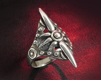 May include: A silver ring with a detailed design featuring a stylized sun with rays and a central circle. The ring has a wide band and a pointed design on the top.