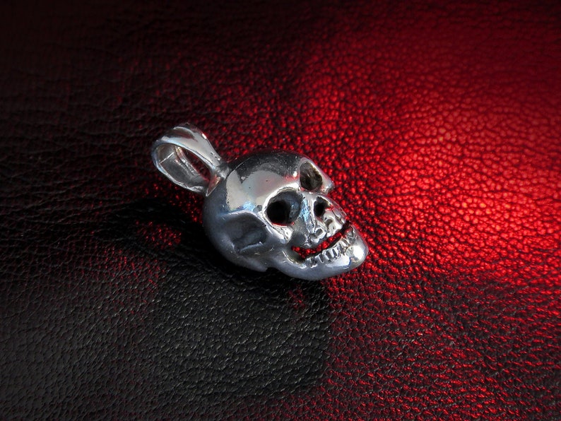 Tiny sterling silver skull pendant resting on textured black leather, illuminated by red accent lighting. The polished surface reflects light across detailed carved features—deep hollow eye sockets, a defined nasal cavity, and precisely articulated teeth. A small, sturdy bail loop at the top hints at versatile threading options for chains or cords, while the unplated finish showcases the authentic “925” silver’s natural luster and artisanal craftsmanship.