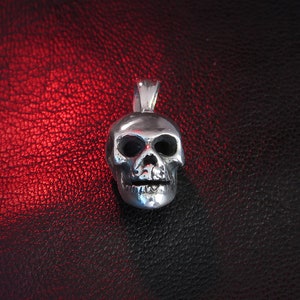 Front-on shot of a miniature hallmarked “925” skull pendant in solid silver, placed on a black leather surface with a subtle red glow. The image captures the pendant’s realistic anatomical proportions—arched brow ridges, sculpted cheekbones, and individually carved teeth. Reflections on the polished metal finish draw attention to the fine craftsmanship and the pendant’s lightweight, wearable scale.