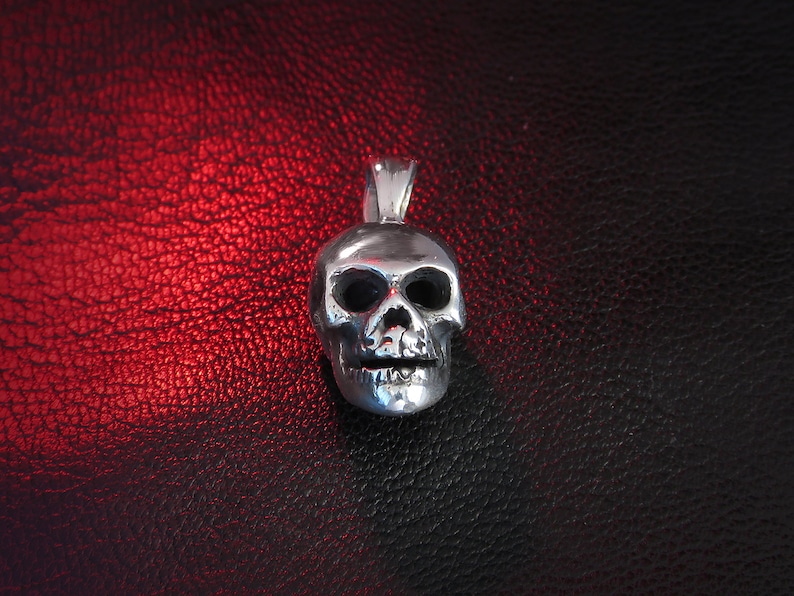 Front-on shot of a miniature hallmarked “925” skull pendant in solid silver, placed on a black leather surface with a subtle red glow. The image captures the pendant’s realistic anatomical proportions—arched brow ridges, sculpted cheekbones, and individually carved teeth. Reflections on the polished metal finish draw attention to the fine craftsmanship and the pendant’s lightweight, wearable scale.