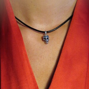Model wearing the miniature sterling silver skull pendant on a slim black leather cord, styled against a deep red blouse. The image demonstrates scale and versatility, showing how the tiny 3.8 g pendant sits comfortably at the collarbone. The unisex gothic-punk design pairs seamlessly with contemporary fashion, illustrating the piece’s statement-making yet understated appeal.
