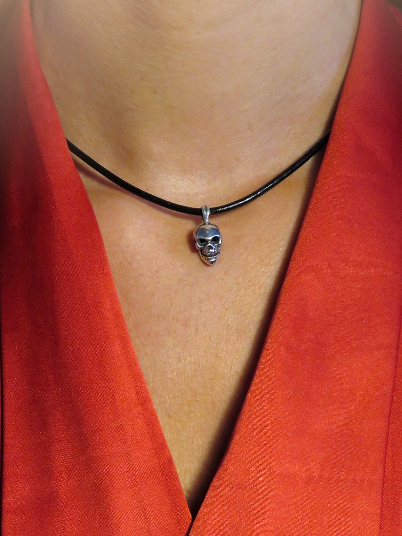Model wearing the miniature sterling silver skull pendant on a slim black leather cord, styled against a deep red blouse. The image demonstrates scale and versatility, showing how the tiny 3.8 g pendant sits comfortably at the collarbone. The unisex gothic-punk design pairs seamlessly with contemporary fashion, illustrating the piece’s statement-making yet understated appeal.