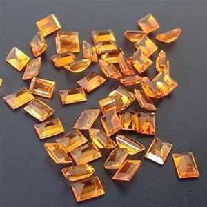 May include: A scattering of orange-yellow gemstones, cut into various shapes such as squares and rectangles. The gems are displayed on a dark surface, reflecting light and showcasing their internal facets.