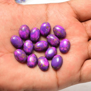 Könnte beinhalten: Eine Sammlung von 15 ovalen Cabochons in Lila und Gold, wahrscheinlich Türkis, in einer Hand. Die Steine haben ein marmoriertes Muster und sind etwa 2,5 cm lang.