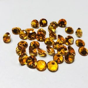 May include: A collection of round, faceted, golden-amber gemstones. The gemstones are scattered on a white surface.