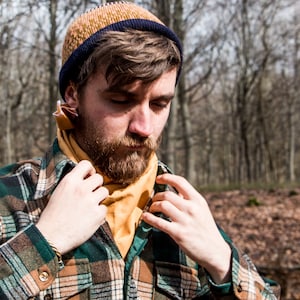 Puede incluir: Un hombre con una camisa de franela a cuadros verde y marrón con una bufanda amarilla en el cuello y una boina de punto marrón y azul. Está de pie en un bosque.