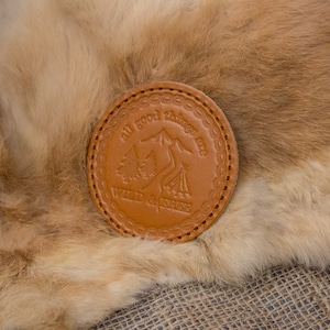 May include: A circular, tan leather coaster featuring the text "all good things are WILD & FREE" and a mountain and tent design. The coaster is stitched around the edge and sits on a fur surface.