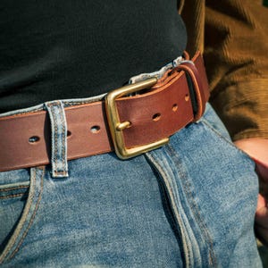 May include: A brown leather belt with a brass buckle is worn with blue jeans. The belt has a rectangular brass buckle and several holes for adjustment. The jeans are a light blue denim.