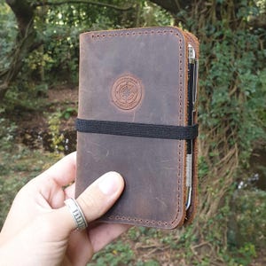 May include: A brown leather notebook with a black elastic band and a pen holder. The notebook has a compass logo with the text 'JOURNEYMAN HANDCRAFTS'.