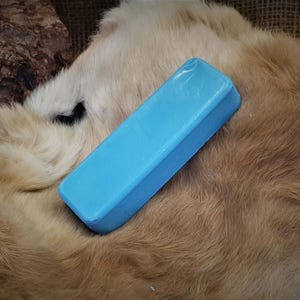 Puede incluir: Una barra rectangular azul claro de jabón descansa sobre una superficie texturizada de color marrón claro. El jabón tiene un aspecto liso y ligeramente brillante. El fondo presenta una textura natural similar a la piel.