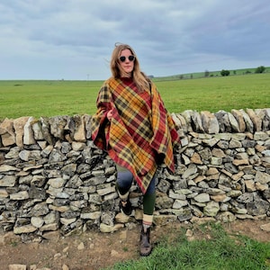 Könnte beinhalten: Ein karierter Poncho in Rot-, Gelb- und Brauntönen. Der Poncho wird über Leggings und Stiefeln getragen. Die Person steht neben einer Steinmauer auf einem Feld.