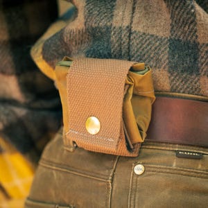 Op de afbeelding: Een kleine, bruin canvas tasje met een gouden knoopsluiting, bevestigd aan een riem. Het tasje is gemaakt van een duurzaam materiaal en heeft een eenvoudig, functioneel ontwerp. De achtergrond omvat een geruit overhemd en een bruine broek.