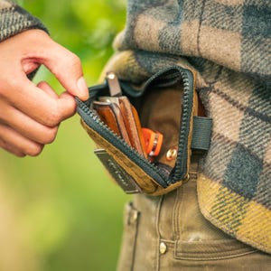 May include: A brown and green zippered pouch attached to a person's clothing. The pouch is open, revealing a brown leather wallet, an orange knife, and other items. The person's hand is pulling the zipper open.