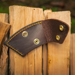 May include: A brown leather sheath with yellow stitching and brass eyelets. The curved sheath is designed for a knife or tool. The background is made of wooden planks.