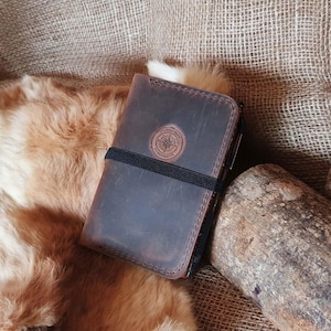 May include: A dark brown leather notebook with a black elastic band closure. The notebook has a compass and the words 'TOGETHER WE ROAM' and 'HANDCRAFTED' embossed on the front cover.