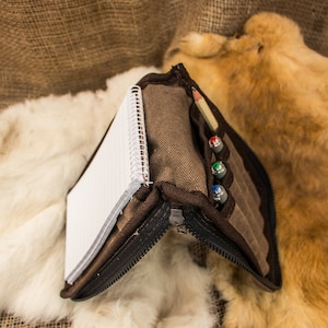 May include: A brown canvas zippered pouch with a notebook and pens inside. The pouch is open and the notebook is visible. The pouch is sitting on a white fur surface.