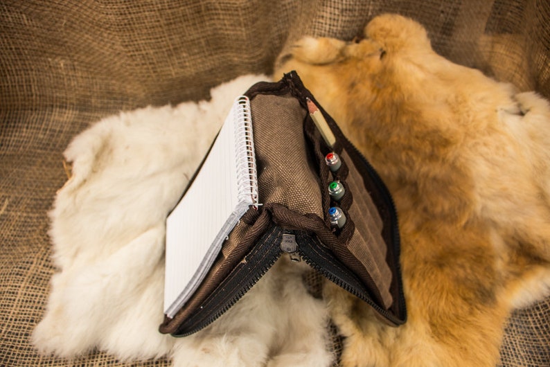May include: A brown canvas zippered pouch with a notebook and pens inside. The pouch is open and the notebook is visible. The pouch is sitting on a white fur surface.