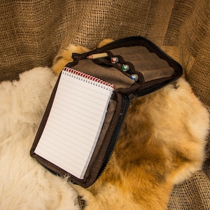 May include: A brown leather pencil case with a zipper closure, containing a notebook and pencils. The case is lying on a white and brown fur surface.
