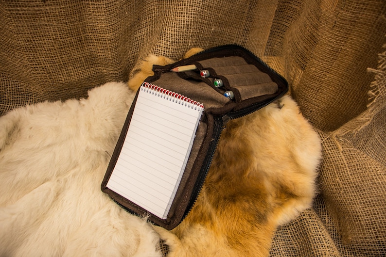 May include: A brown leather pencil case with a zipper closure, containing a notebook and pencils. The case is lying on a white and brown fur surface.