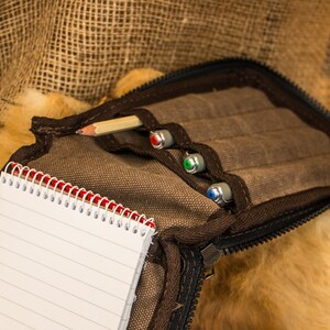 May include: A brown canvas pencil case with a zipper closure. The case is open and contains a pencil, three pens, and a small notebook with red and white spiral binding.