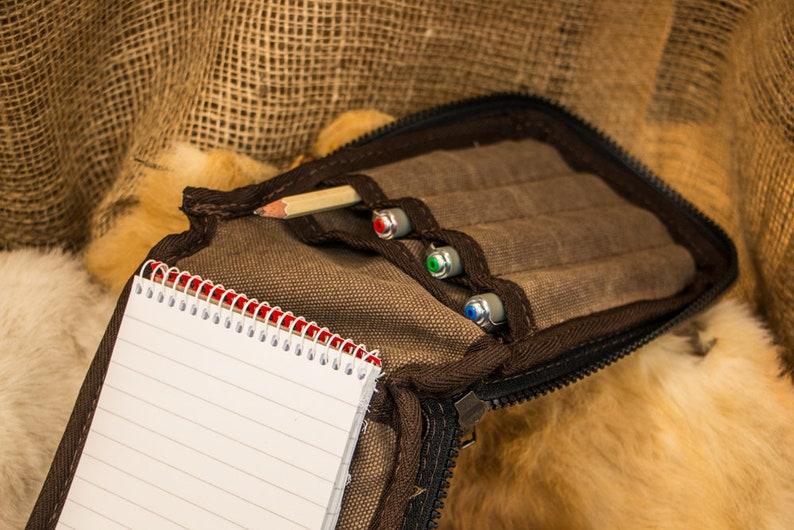 May include: A brown canvas pencil case with a zipper closure. The case is open and contains a pencil, three pens, and a small notebook with red and white spiral binding.