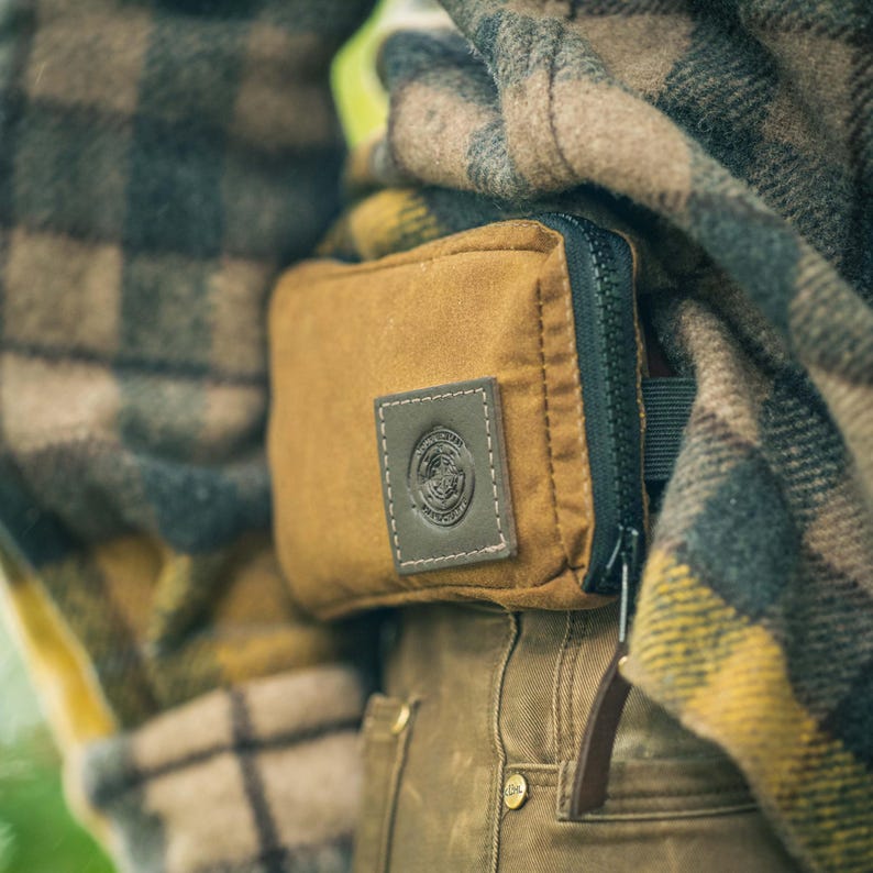 May include: A close-up of a brown canvas pouch with a black zipper and a dark green leather patch. The patch has a circular emblem and white stitching. The pouch is attached to a person wearing a plaid jacket and brown pants.