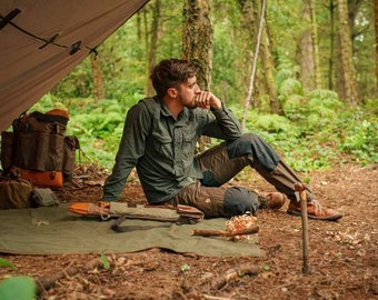 May include: A person sits under a tarp in a forest, wearing a green shirt and brown pants. A backpack, orange bag, and other gear are nearby. A knife sheath and a green mat are on the ground, suggesting outdoor activities.
