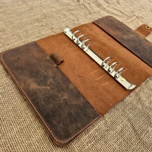 Può includere: Raccoglitore in pelle marrone con finitura invecchiata aperto. L'interno è marrone chiaro con un meccanismo ad anelli in metallo argentato. Una tasca è visibile sul lato sinistro. Il raccoglitore è aperto, rivelando l'interno.