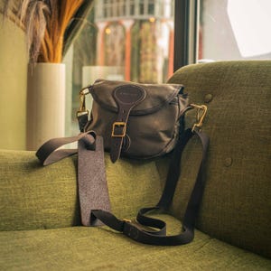 May include: A dark green canvas shoulder bag with a leather strap and buckle closure. The bag has a long, adjustable black strap and gold-coloured metal hardware. The bag is on a green sofa.