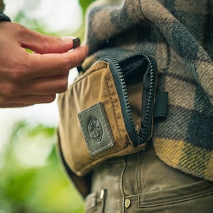 May include: A brown canvas bag with a black zip and a leather patch, attached to a plaid jacket. The zip is partly open, and a hand is reaching towards it. The jacket has a green, brown, and yellow plaid pattern.