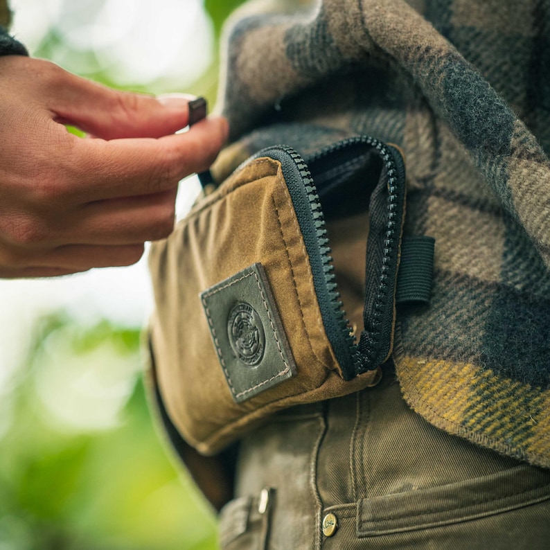 May include: A close-up of a brown canvas bag with a black zipper and a leather patch. The bag is attached to a plaid jacket. The zipper is partially open, and a hand is reaching towards it.