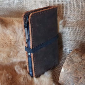 May include: A brown leather notebook with a black elastic band closure and a black pen inserted in the side pocket. The notebook is on a brown fur surface.