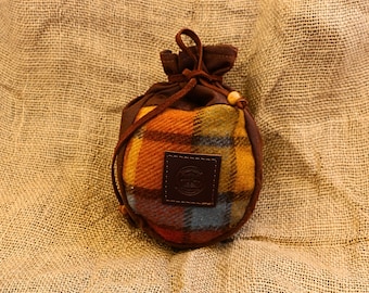 May include: A small, round drawstring pouch with a plaid pattern in shades of orange, yellow, blue, and red. The pouch has a brown leather patch and a brown drawstring closure. The background is a textured, tan fabric.