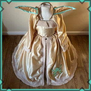 Puede incluir: Un vestido de sat&eacute;n dorado con ribete blanco esponjoso y una aplicaci&oacute;n de rana verde. El vestido tiene una diadema a juego con orejas grandes de color azul claro.