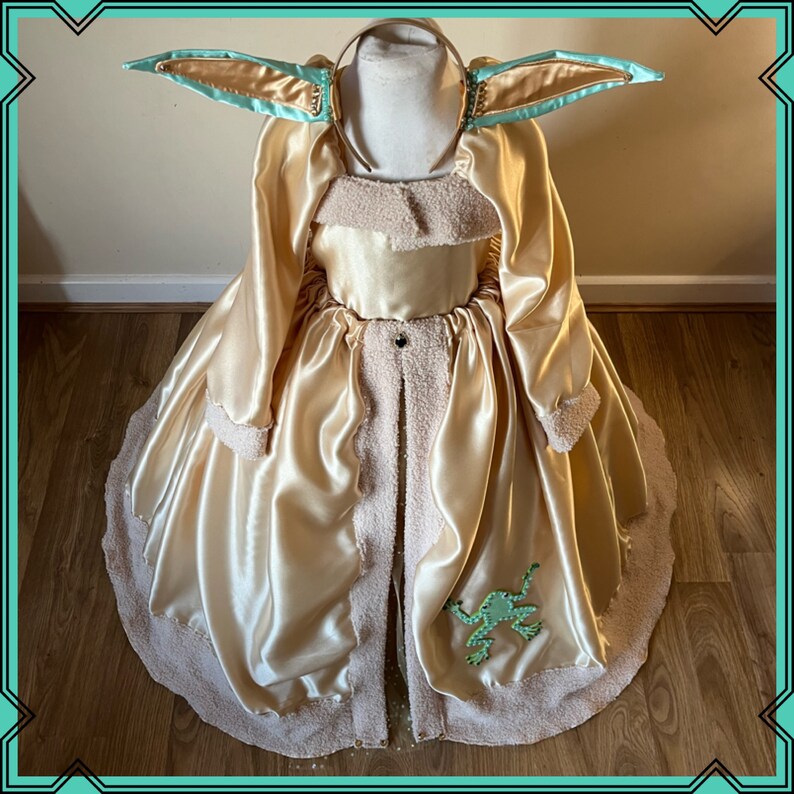 Puede incluir: Un vestido de sat&eacute;n dorado con ribete blanco esponjoso y una aplicaci&oacute;n de rana verde. El vestido tiene una diadema a juego con orejas grandes de color azul claro.