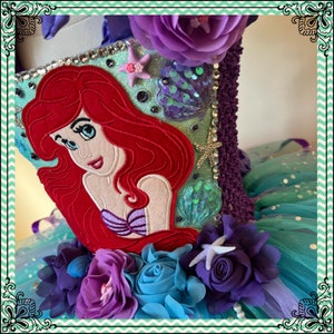 Puede incluir: Un tut&uacute; de tul turquesa, morado y verde con una Ariel roja bordada de La Sirenita. El tut&uacute; est&aacute; decorado con flores moradas, azules y turquesas, y una estrella de mar.
