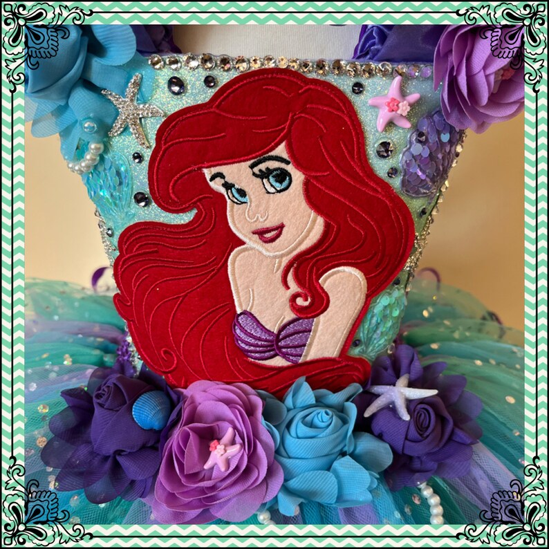 Puede incluir: Un tut&uacute; de tul turquesa con una Ariel bordada en rojo y blanco de La Sirenita. El tut&uacute; est&aacute; decorado con flores moradas, azules y blancas, conchas marinas y estrellas de mar.