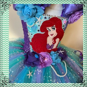 Puede incluir: Un tut&uacute; de tul turquesa con un adorno de flores moradas y azules. El corpi&ntilde;o est&aacute; decorado con una aplicaci&oacute;n de Ariel de fieltro rojo, pedrer&iacute;a y una hebra de perlas.