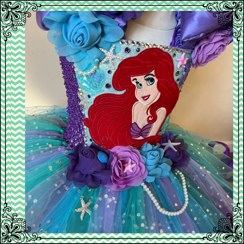 Puede incluir: Un tut&uacute; de tul turquesa con un adorno de flores moradas y azules. El corpi&ntilde;o est&aacute; decorado con una aplicaci&oacute;n de Ariel de fieltro rojo, pedrer&iacute;a y una hebra de perlas.
